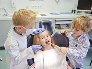 Мальчик переоделся в доктора осматривает девочку на стоматологическом кресле. Ему помогает еще один малыш
